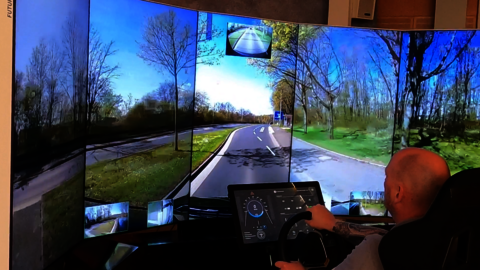 A man is sitting in front of 5 large portrait screens with a steering wheel in his hand. On the screens you can see a road and green meadows next to the road.